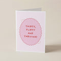 A folded greeting card standing upright on a light beige surface. The card is white with a pink scalloped oval in the center containing the text 'THIRTY, FLIRTY AND THRIVING!' in pink lettering.