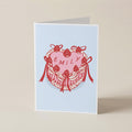 A folded greeting card standing upright on a light beige surface. The card has a pale blue background with a central illustration of a pink heart layered over a red floral wreath with bows, and the name 'EMILY' printed across the heart.