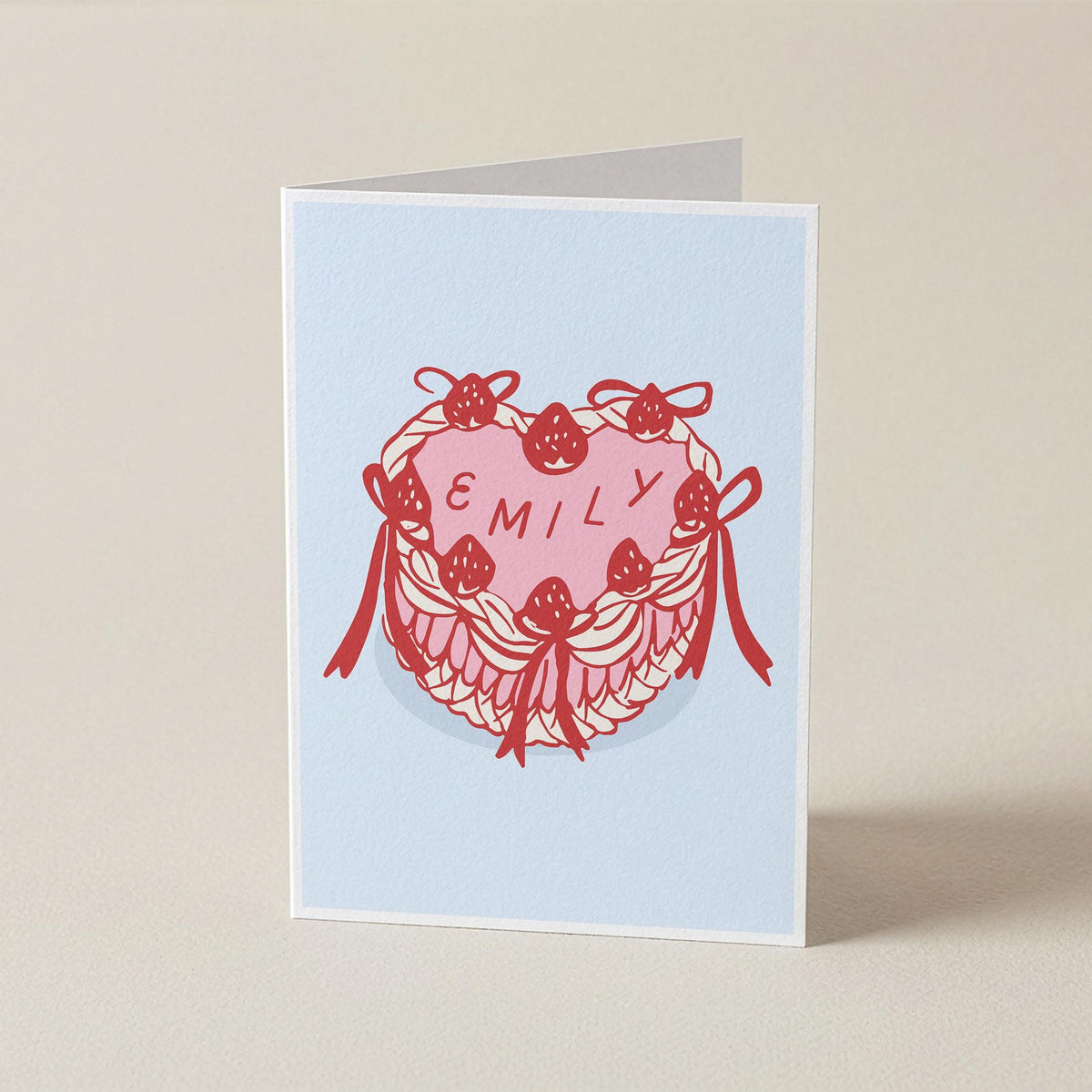 A folded greeting card standing upright on a light beige surface. The card has a pale blue background with a central illustration of a pink heart layered over a red floral wreath with bows, and the name 'EMILY' printed across the heart.