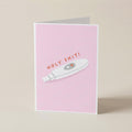 A folded greeting card standing upright on a light beige surface. The card has a pink front with an illustration of a white pregnancy test and the text 'HOLY SHIT!' in orange-red lettering.