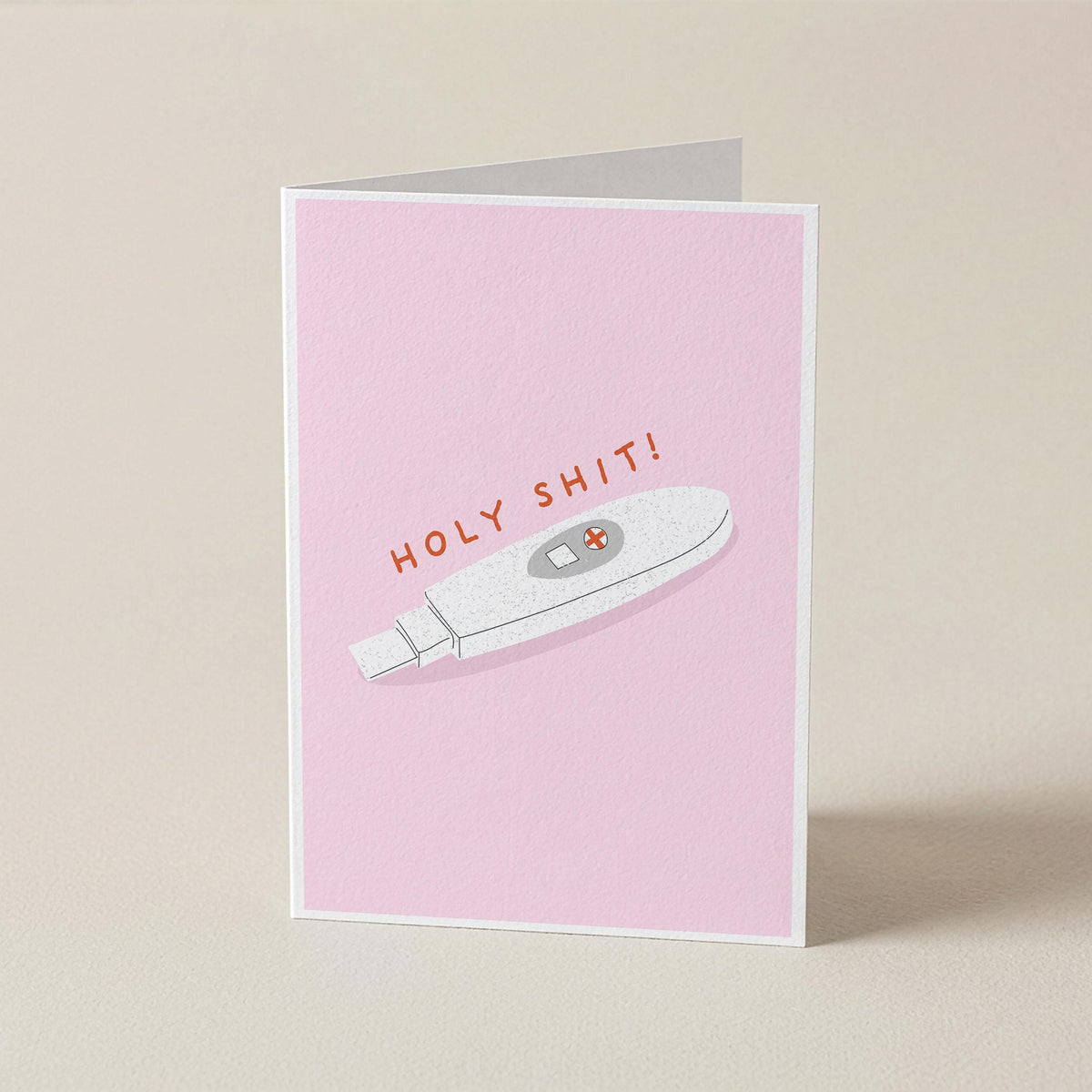 A folded greeting card standing upright on a light beige surface. The card has a pink front with an illustration of a white pregnancy test and the text 'HOLY SHIT!' in orange-red lettering.