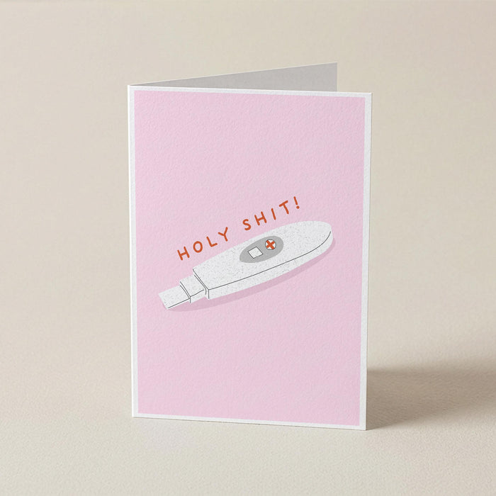 A folded greeting card standing upright on a light beige surface. The card has a pink front with an illustration of a white pregnancy test and the text 'HOLY SHIT!' in orange-red lettering.