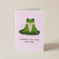 A folded greeting card standing upright on a light beige surface. The card has a pale pink background with a cartoon green frog sitting on a green lily pad and the text 'CONGRATS ON YOUR NEW PAD!' below.