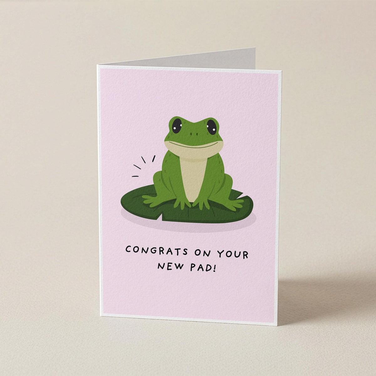 A folded greeting card standing upright on a light beige surface. The card has a pale pink background with a cartoon green frog sitting on a green lily pad and the text 'CONGRATS ON YOUR NEW PAD!' below.