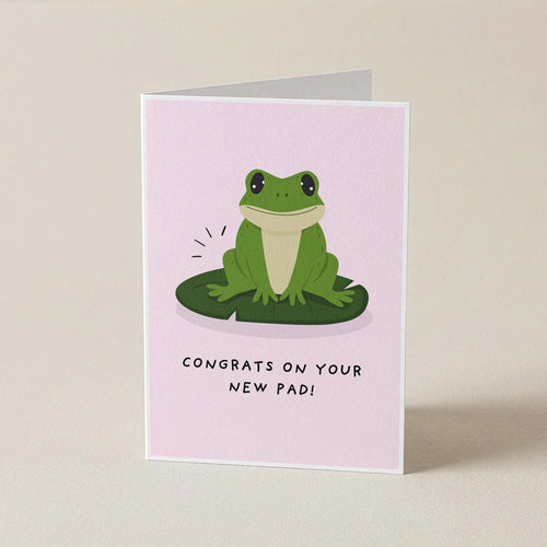 A folded greeting card standing upright on a light beige surface. The card has a pale pink background with a cartoon green frog sitting on a green lily pad and the text 'CONGRATS ON YOUR NEW PAD!' below.