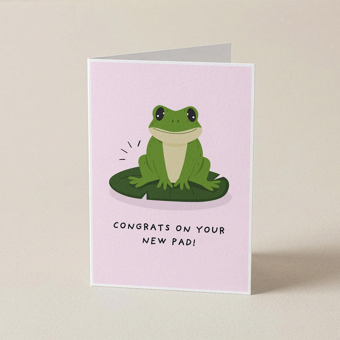 A folded greeting card standing upright on a light beige surface. The card has a pale pink background with a cartoon green frog sitting on a green lily pad and the text 'CONGRATS ON YOUR NEW PAD!' below.