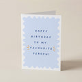 A folded greeting card standing upright on a light beige surface. The front has a white border with a pale blue scalloped shape in the center and the text 'HAPPY BIRTHDAY TO MY FAVOURITE PERSON!' in dark blue, with small orange burst accents.