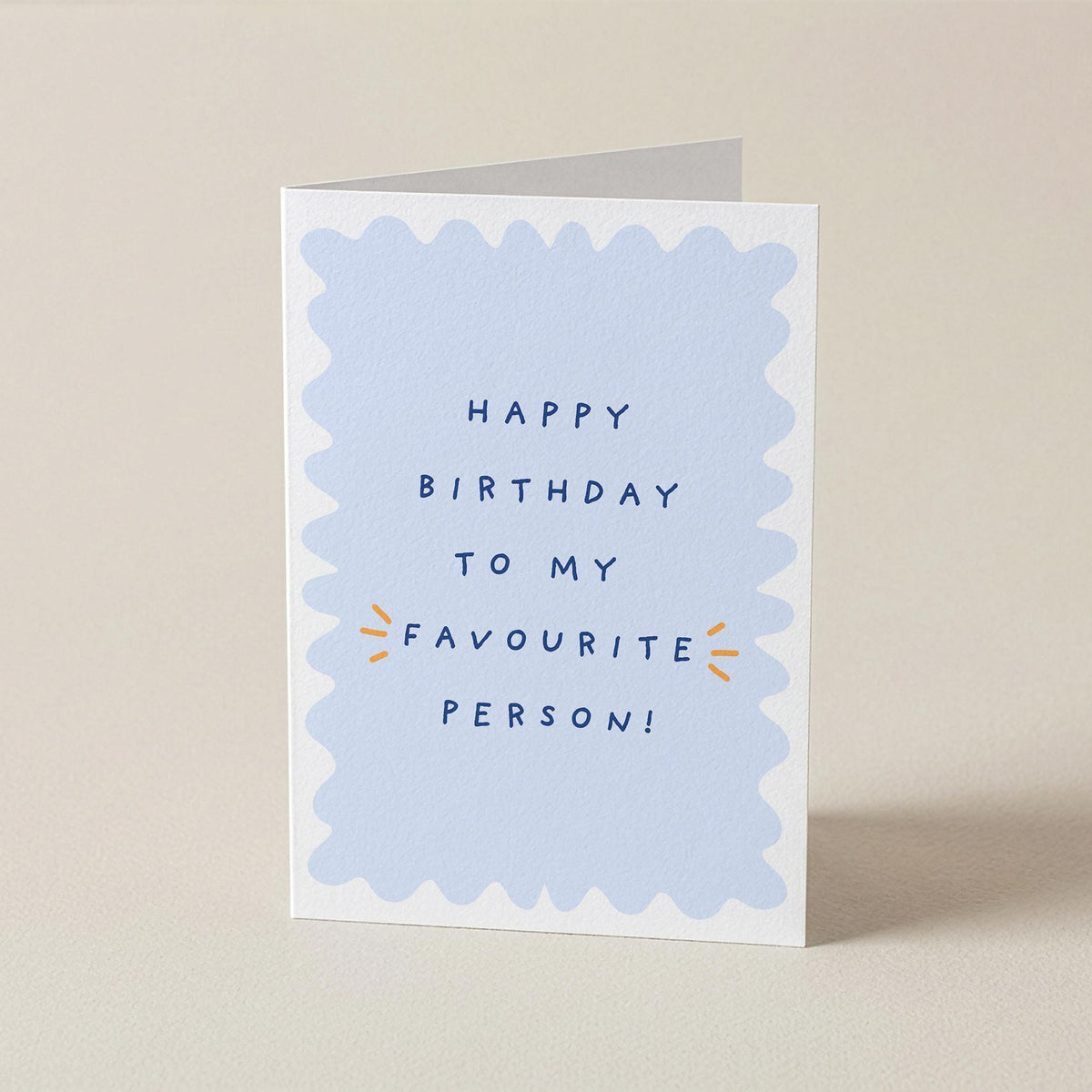 A folded greeting card standing upright on a light beige surface. The front has a white border with a pale blue scalloped shape in the center and the text 'HAPPY BIRTHDAY TO MY FAVOURITE PERSON!' in dark blue, with small orange burst accents.