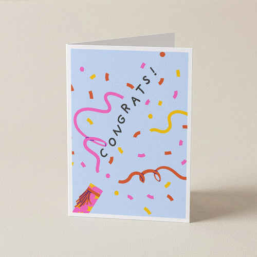 A folded greeting card standing upright on a light beige surface. The card has a pale blue background with the word 'CONGRATS!' in black, surrounded by colorful confetti, streamers, and a small party hat illustration.