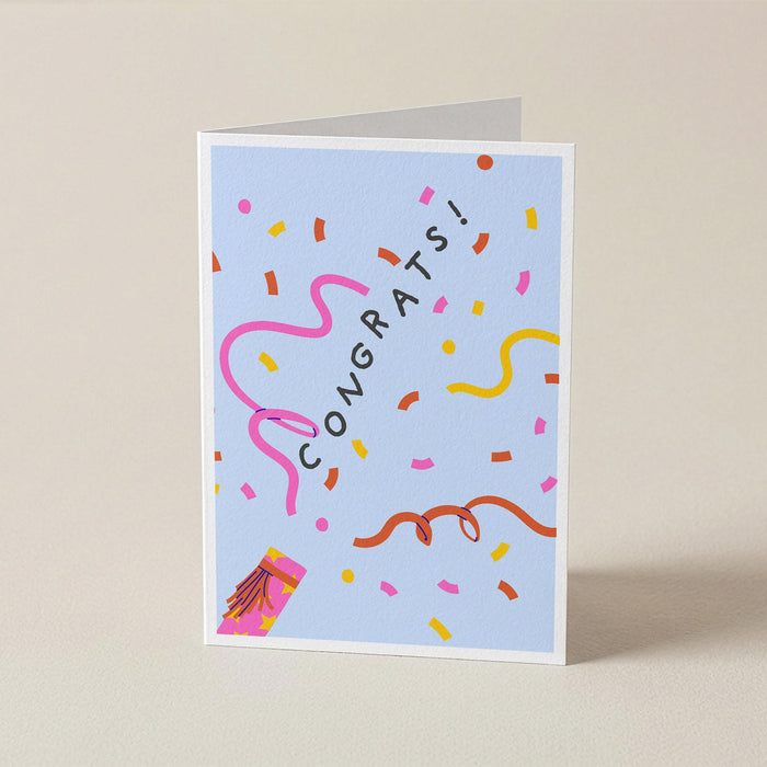 A folded greeting card standing upright on a light beige surface. The card has a pale blue background with the word 'CONGRATS!' in black, surrounded by colorful confetti, streamers, and a small party hat illustration.