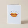 A folded greeting card standing upright on a light beige surface. The card has a white background with a cartoon hot dog in a bun and the text 'YOU CLEVER SAUSAGE!' arched above it.