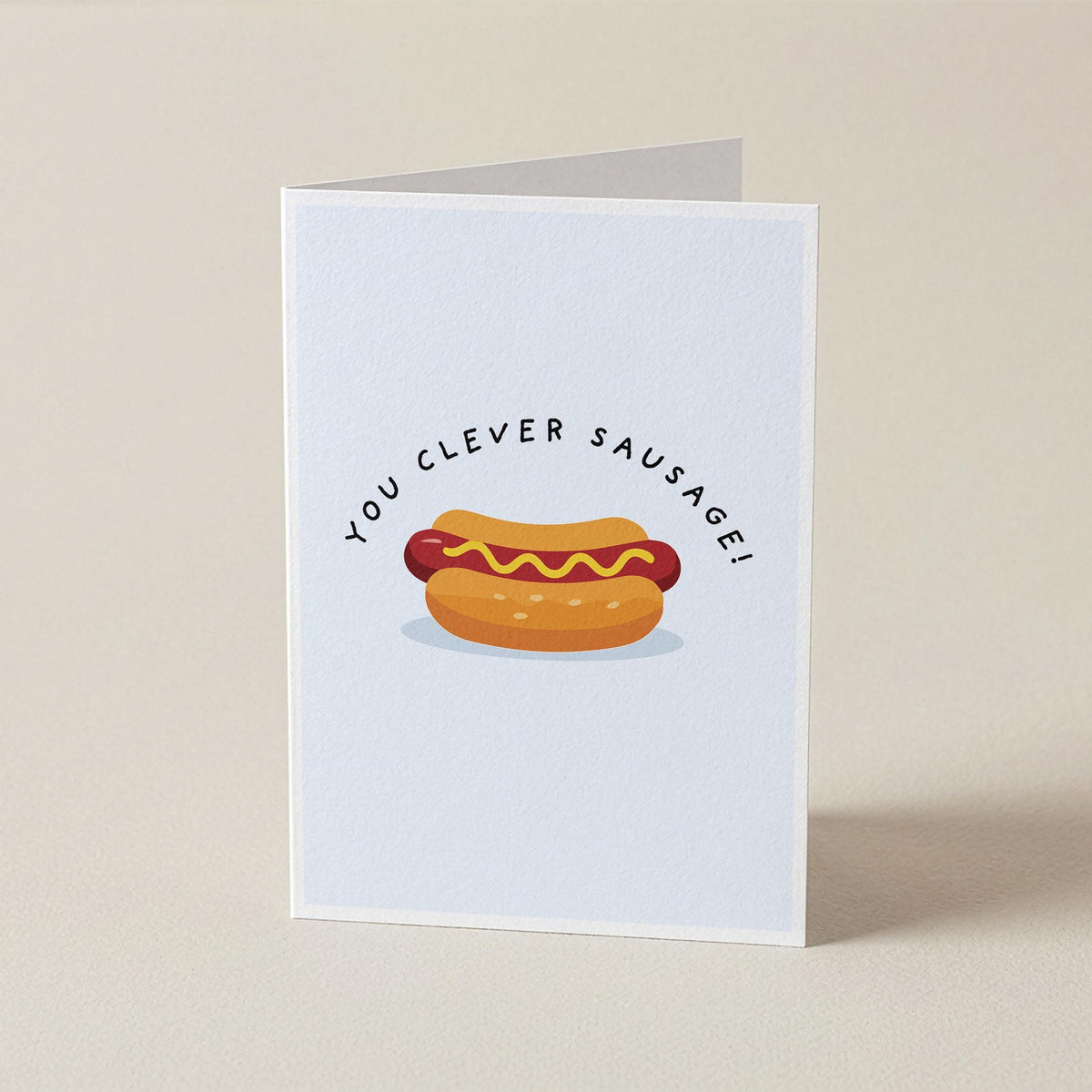 A folded greeting card standing upright on a light beige surface. The card has a white background with a cartoon hot dog in a bun and the text 'YOU CLEVER SAUSAGE!' arched above it.