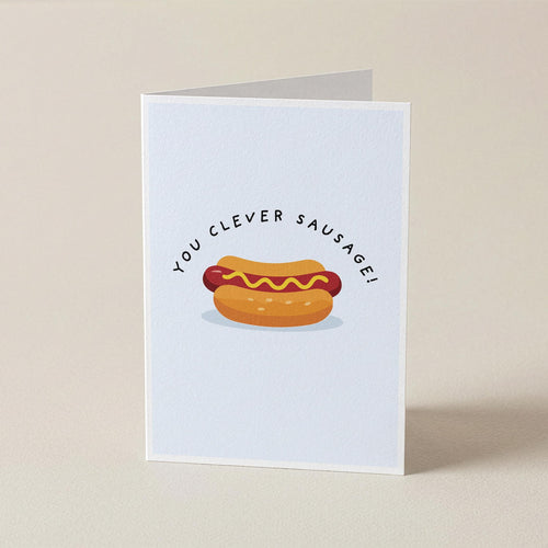 A folded greeting card standing upright on a light beige surface. The card has a white background with a cartoon hot dog in a bun and the text 'YOU CLEVER SAUSAGE!' arched above it.