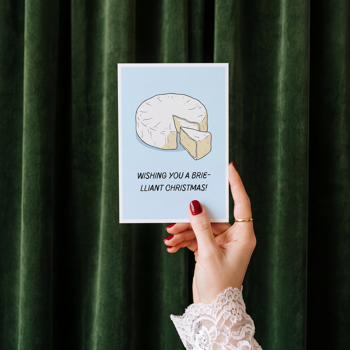 A hand with red nail polish and a white lace cuff holds a light blue greeting card featuring an illustration of a sliced brie cheese and the text 'WISHING YOU A BRIE-LLIANT CHRISTMAS!'. The background is a dark green velvet curtain.