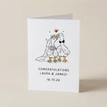 A white greeting card standing upright on a beige surface, featuring a hand-drawn illustration of two wedding birds with a small red heart above them and the text 'CONGRATULATIONS LAURA & JAMES! 16.10.