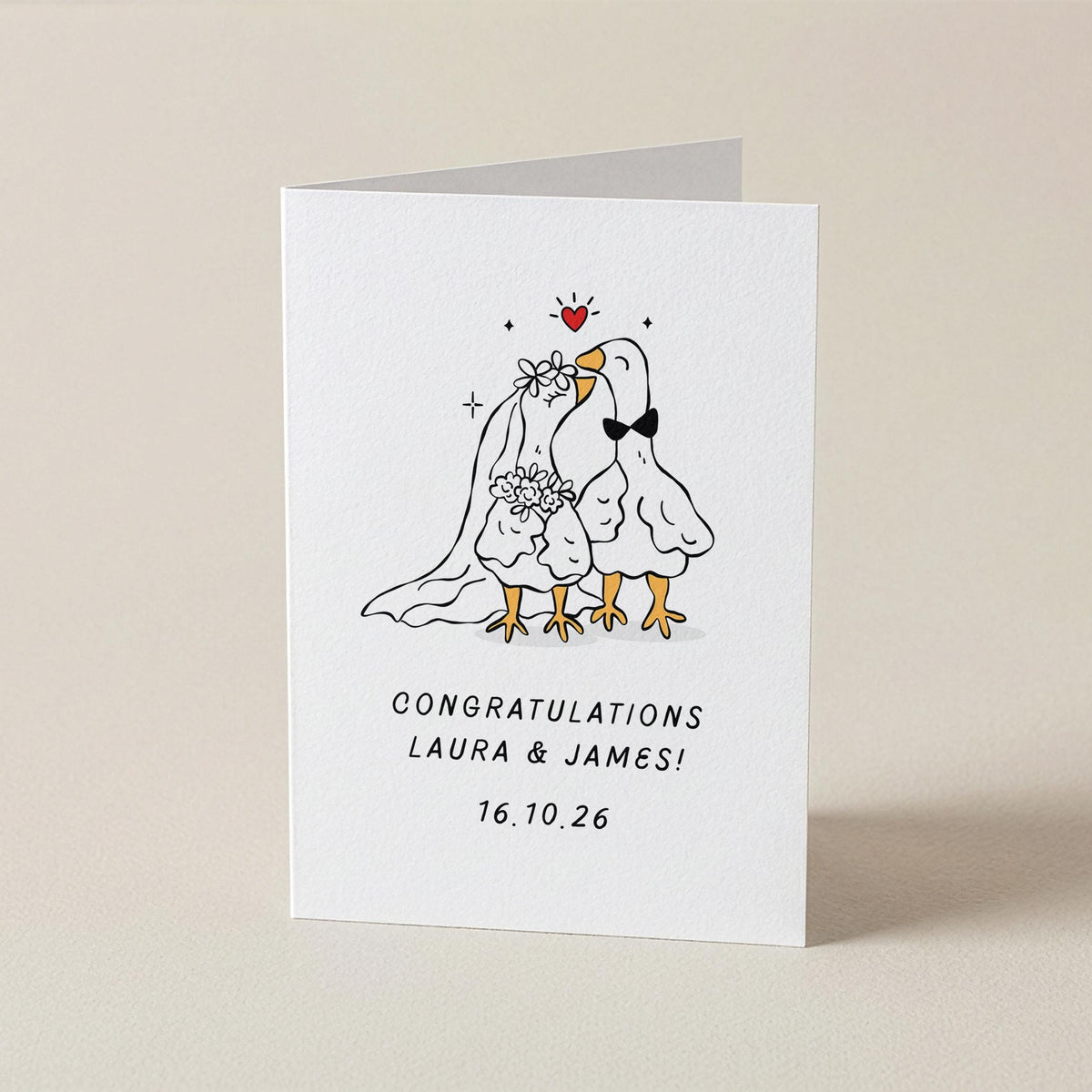 A white greeting card standing upright on a beige surface, featuring a hand-drawn illustration of two wedding birds with a small red heart above them and the text 'CONGRATULATIONS LAURA & JAMES! 16.10.