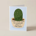 A folded greeting card standing upright on a light beige surface. The card features a simple illustration of a green cactus in a beige pot with the text 'HOPE YOUR NEW NEIGHBOURS AREN'T PRICKS!' on a pale blue background.