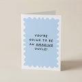 A folded greeting card standing upright on a light beige surface. The card has a white border with a light blue scalloped inner shape and black text that reads, 'YOU'RE GOING TO BE AN AMAZING UNCLE!' with 'AMAZING' underlined.