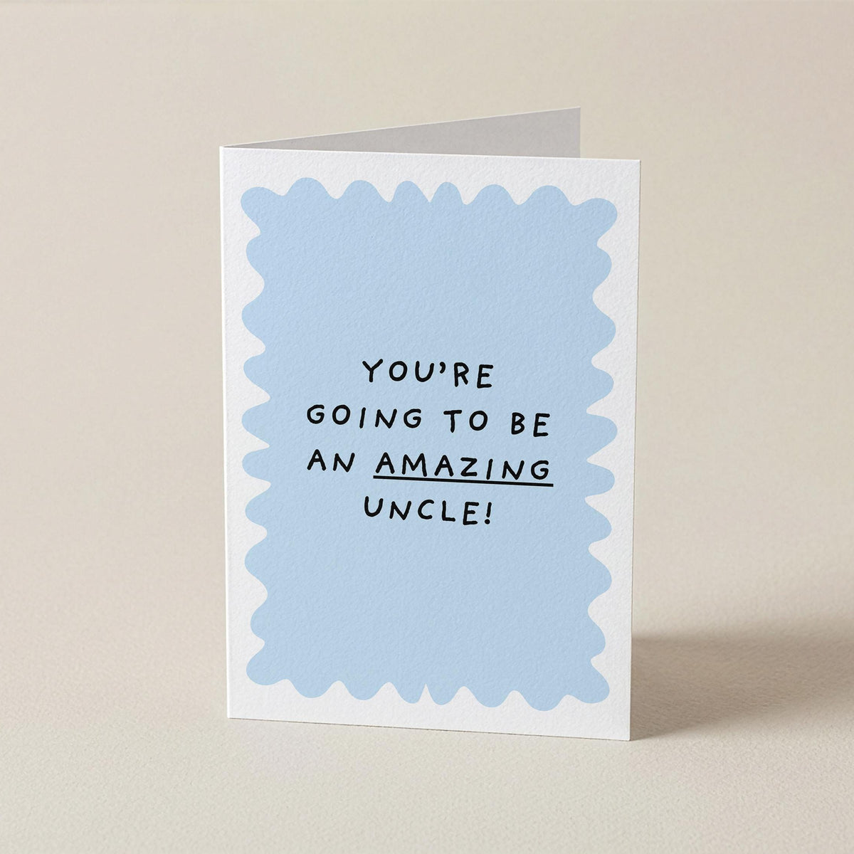 A folded greeting card standing upright on a light beige surface. The card has a white border with a light blue scalloped inner shape and black text that reads, 'YOU'RE GOING TO BE AN AMAZING UNCLE!' with 'AMAZING' underlined.