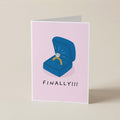 A folded greeting card standing upright on a light beige surface. The card has a pale pink background and features an illustration of an open blue ring box with a yellow ring and a diamond, above the text 'FINALLY!!!' in black.