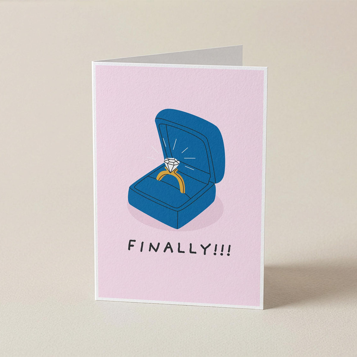A folded greeting card standing upright on a light beige surface. The card has a pale pink background and features an illustration of an open blue ring box with a yellow ring and a diamond, above the text 'FINALLY!!!' in black.