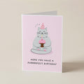 A folded greeting card standing upright on a light beige background. The card has a pale pink front with a cartoon cat wearing a party hat and holding a cupcake, with the text 'HOPE YOU HAVE A PURRRRRECT BIRTHDAY' below.
