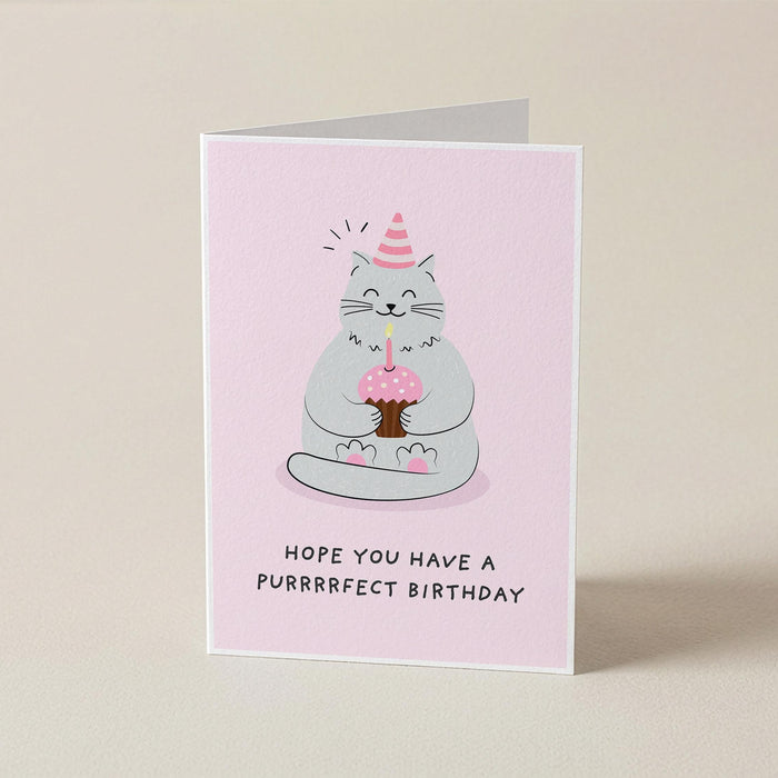 A folded greeting card standing upright on a light beige background. The card has a pale pink front with a cartoon cat wearing a party hat and holding a cupcake, with the text 'HOPE YOU HAVE A PURRRRRECT BIRTHDAY' below.