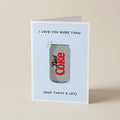 A folded greeting card standing upright on a light beige surface. The card has a pale blue front with a printed illustration of a gray Diet Coke can and the text 'I LOVE YOU MORE THAN (AND THATS A LOT)'.