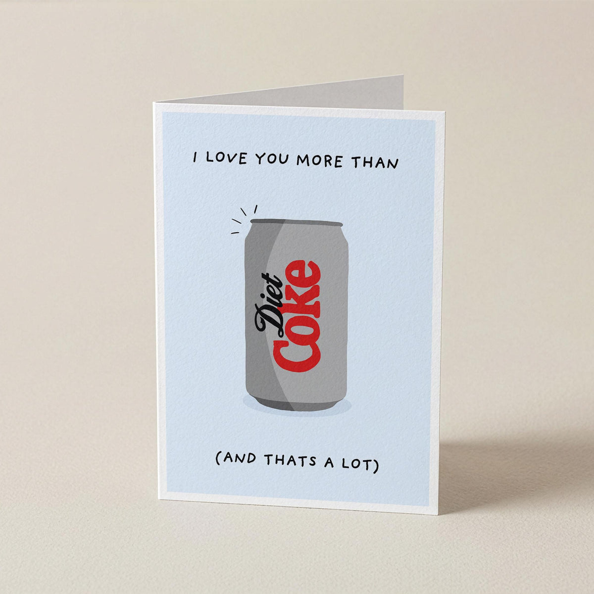 A folded greeting card standing upright on a light beige surface. The card has a pale blue front with a printed illustration of a gray Diet Coke can and the text 'I LOVE YOU MORE THAN (AND THATS A LOT)'.