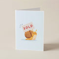 A folded greeting card standing upright on a light beige surface. The card has a white background with a cartoon snail holding a sign that says 'SOLD' in red letters.