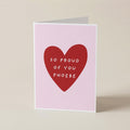 A folded greeting card standing upright on a light beige surface. The card has a pale pink background with a large red heart in the center containing the white text 'SO PROUD OF YOU PHOEBE'.