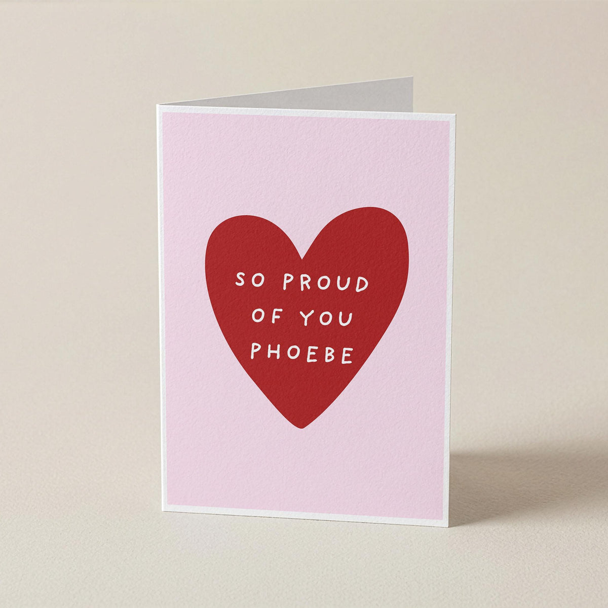 A folded greeting card standing upright on a light beige surface. The card has a pale pink background with a large red heart in the center containing the white text 'SO PROUD OF YOU PHOEBE'.