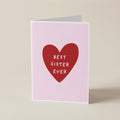 A folded greeting card standing upright on a light beige surface. The card has a pale pink background with a large red heart in the center containing the white text 'BEST SISTER EVER'.