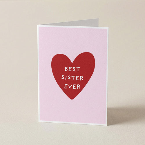 A folded greeting card standing upright on a light beige surface. The card has a pale pink background with a large red heart in the center containing the white text 'BEST SISTER EVER'.