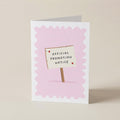 A folded greeting card standing upright on a light beige surface. The card has a white border and a pale pink scalloped background, with a small illustrated sign reading 'OFFICIAL PROMOTION NOTICE' on a stick.