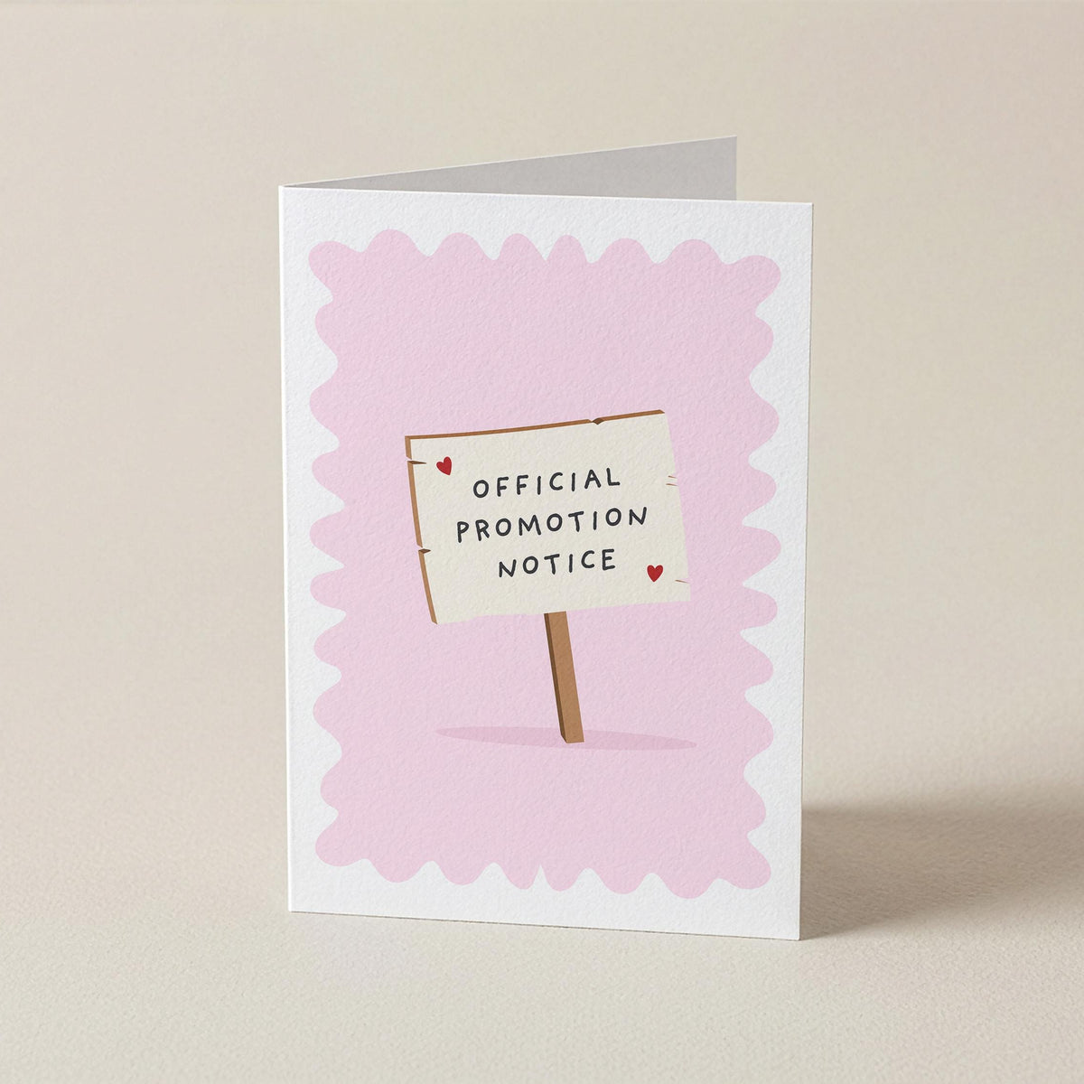 A folded greeting card standing upright on a light beige surface. The card has a white border and a pale pink scalloped background, with a small illustrated sign reading 'OFFICIAL PROMOTION NOTICE' on a stick.