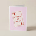 A folded greeting card standing upright on a light beige surface. The card has a pale pink background with a central illustration of a playing card labeled 'QUEEN OF MUMS' in red text and heart symbols.