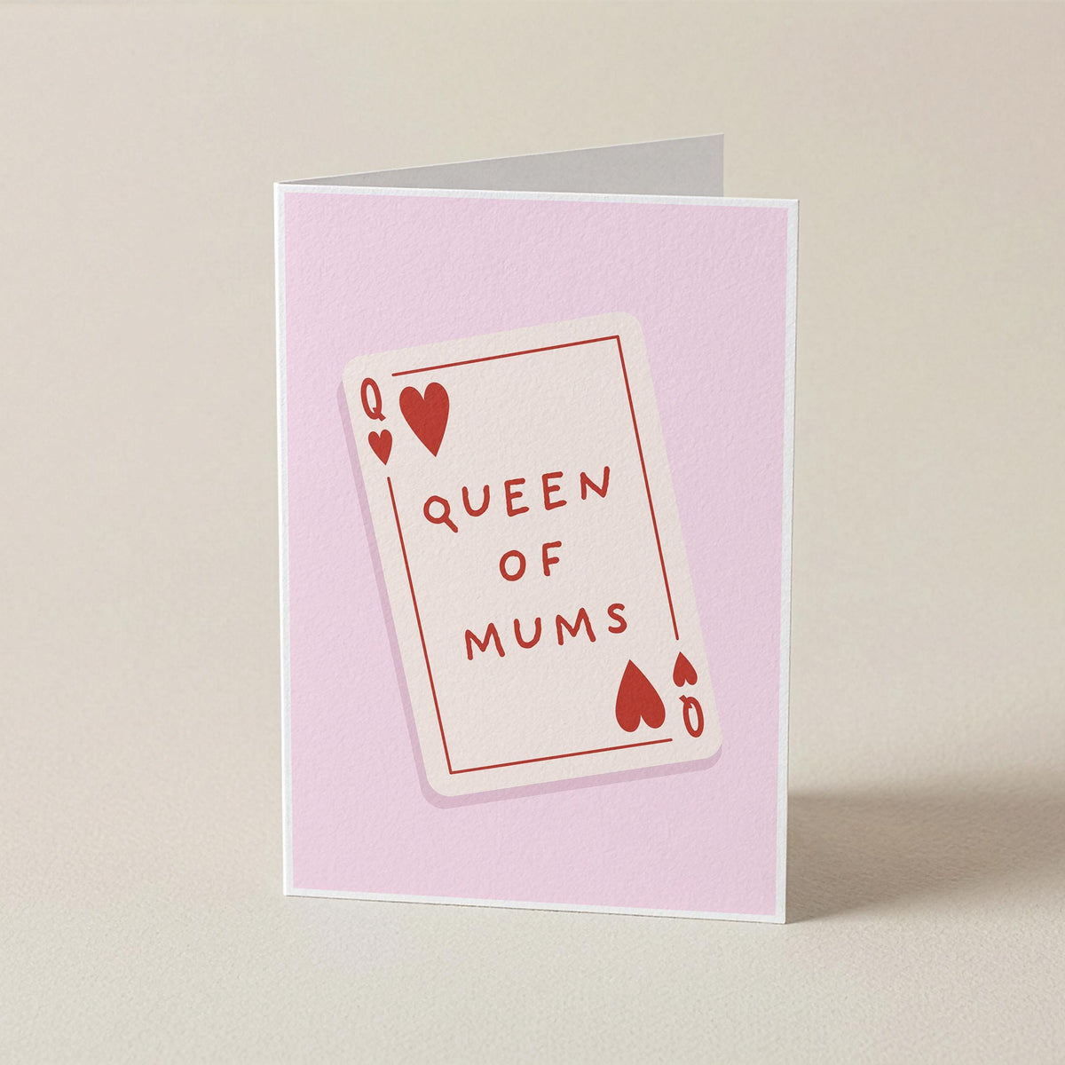 A folded greeting card standing upright on a light beige surface. The card has a pale pink background with a central illustration of a playing card labeled 'QUEEN OF MUMS' in red text and heart symbols.
