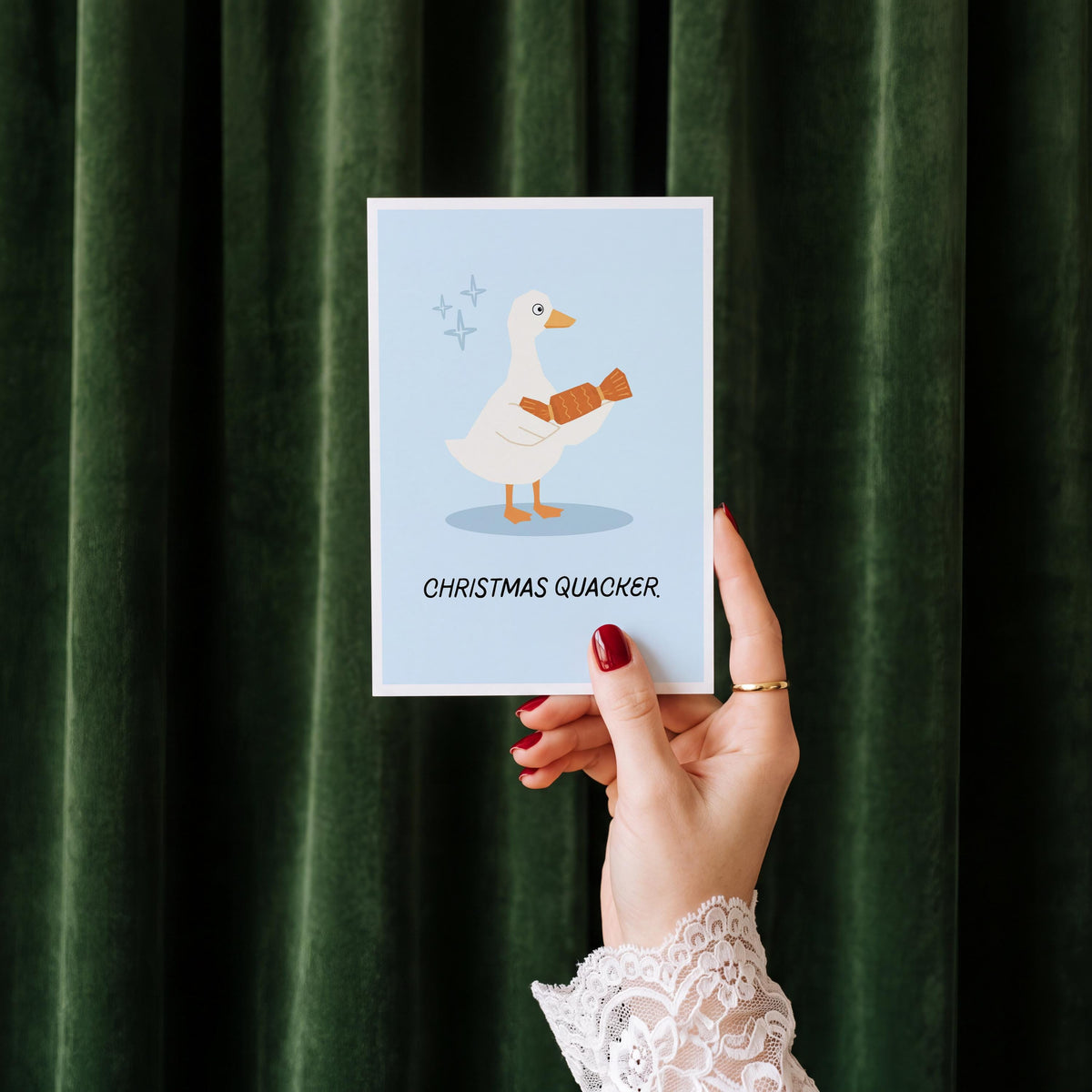 A hand with red nail polish and a white lace cuff holds up a square art print featuring a white Christmas quacker duck with a brown scarf on a light blue background, set against a dark green velvet curtain backdrop.