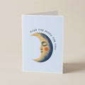 A folded greeting card standing upright on a light beige surface. The card has a pale blue front with an illustration of a crescent moon face and the text 'OVER THE MOON FOR YOU'.