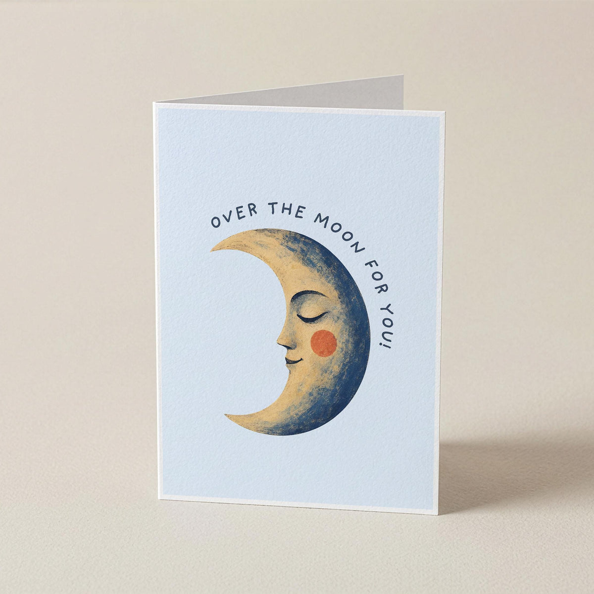A folded greeting card standing upright on a light beige surface. The card has a pale blue front with an illustration of a crescent moon face and the text 'OVER THE MOON FOR YOU'.