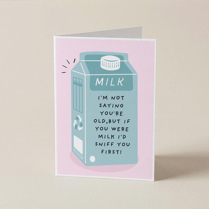 A folded greeting card standing upright on a light beige surface. The card has a pink border and features an illustration of a milk carton in light blue with the text, 'MILK I'M NOT SAYING YOU'RE OLD, BUT IF YOU WERE MILK I'D SNIFF YOU FIRST!'.