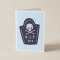 A folded greeting card standing upright on a light beige surface. The card has a pale blue front with a dark navy tombstone graphic featuring a white skull and crossbones and the text 'R.