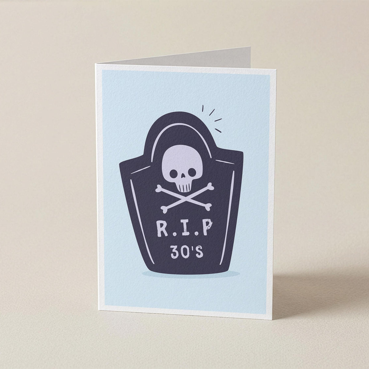 A folded greeting card standing upright on a light beige surface. The card has a pale blue front with a dark navy tombstone graphic featuring a white skull and crossbones and the text 'R.