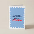 A folded greeting card standing upright on a light beige surface. The card has a white border, a light blue scalloped central panel, the text 'HAVE A RAIL-Y GOOD BIRTHDAY' in dark blue, and a small red train illustration across the middle.