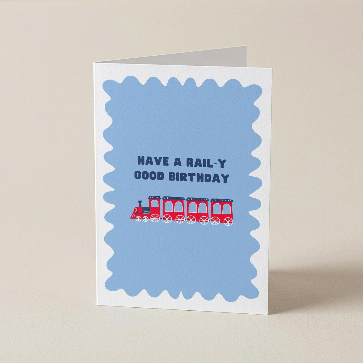 A folded greeting card standing upright on a light beige surface. The card has a white border, a light blue scalloped central panel, the text 'HAVE A RAIL-Y GOOD BIRTHDAY' in dark blue, and a small red train illustration across the middle.