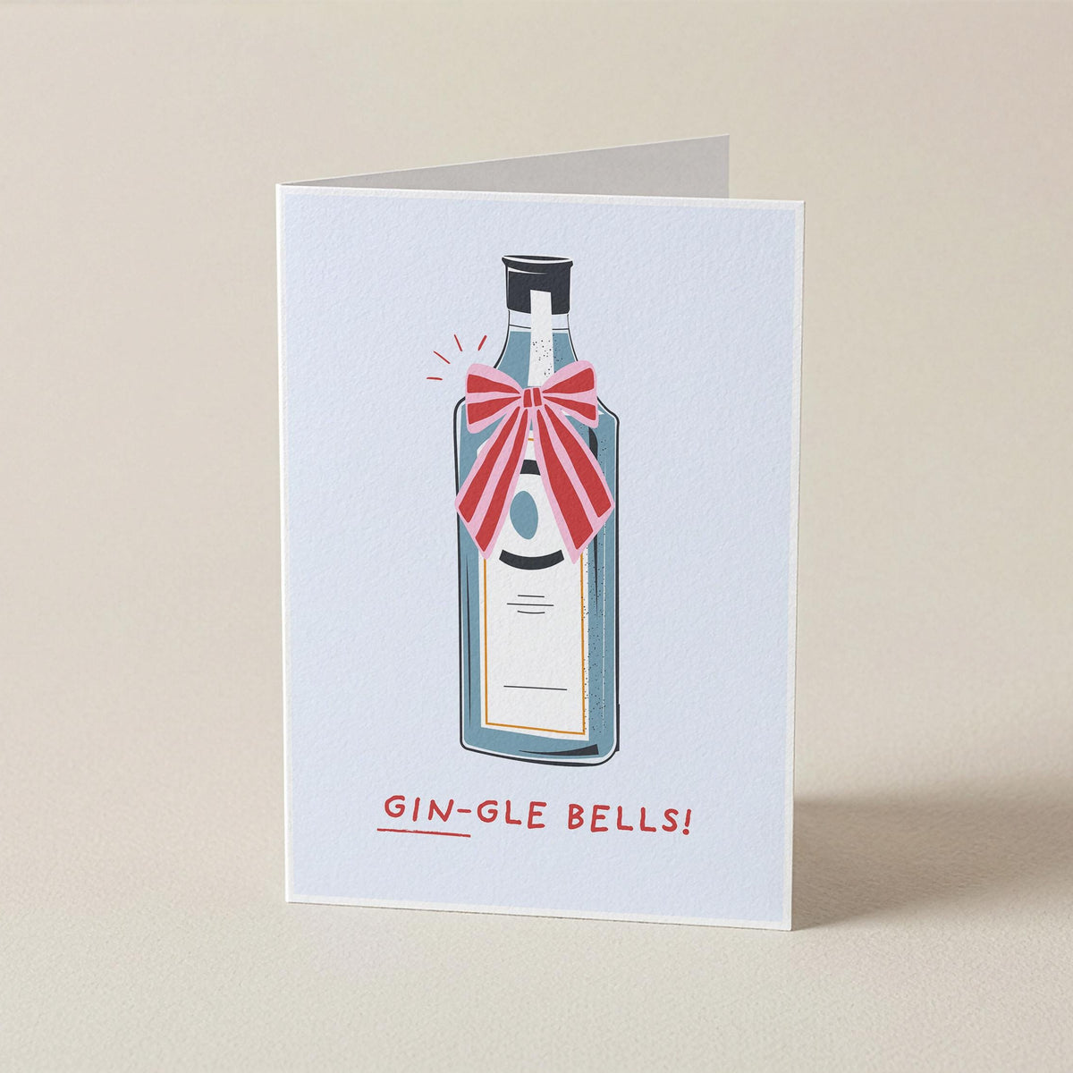 A folded greeting card standing upright on a beige surface. The card features an illustration of a gin bottle with a red striped bow and the text 'GIN-GLE BELLS!' printed below.