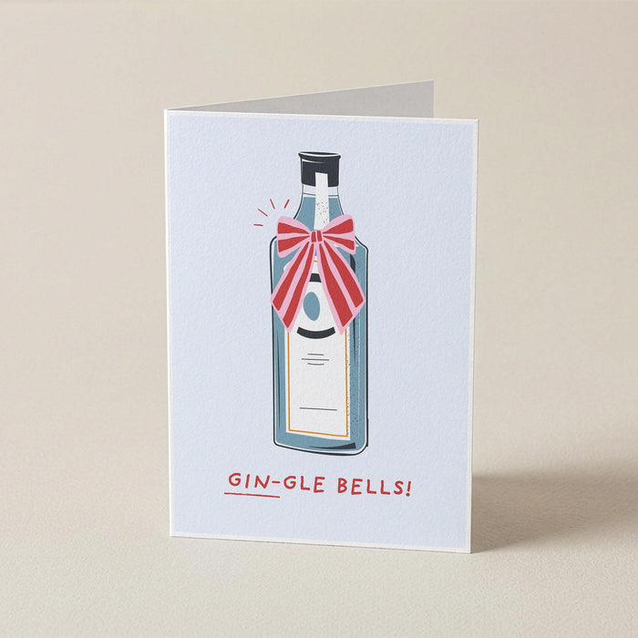 A folded greeting card standing upright on a beige surface. The card features an illustration of a gin bottle with a red striped bow and the text 'GIN-GLE BELLS!' printed below.