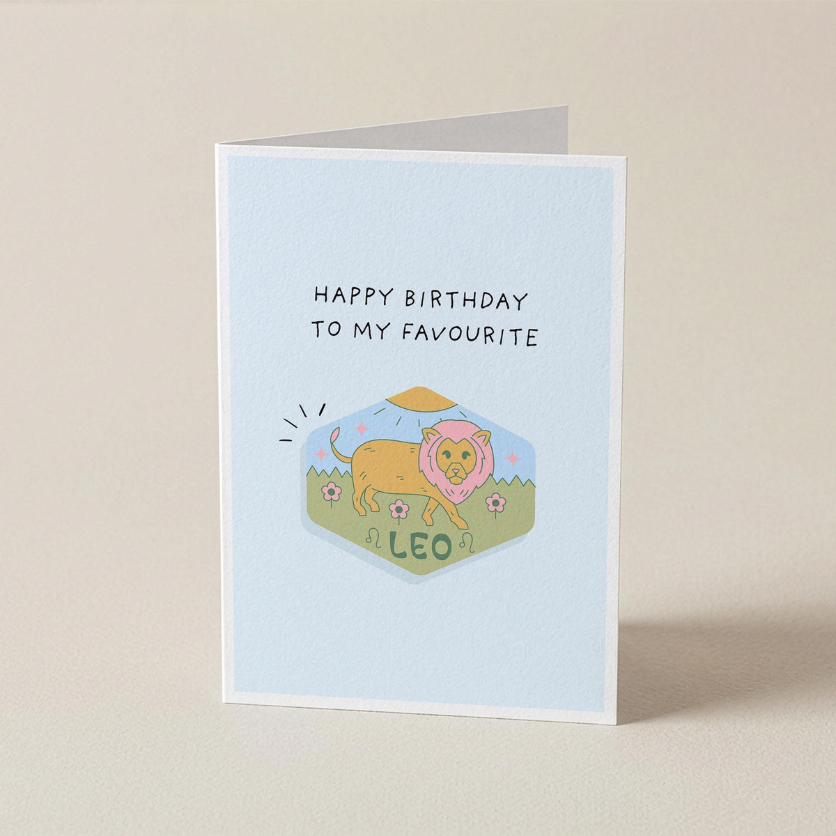 A folded greeting card standing upright on a light beige surface. The card has a pale blue front with the text 'HAPPY BIRTHDAY TO MY FAVOURITE' above a small illustrated lion labeled 'A LEO'.