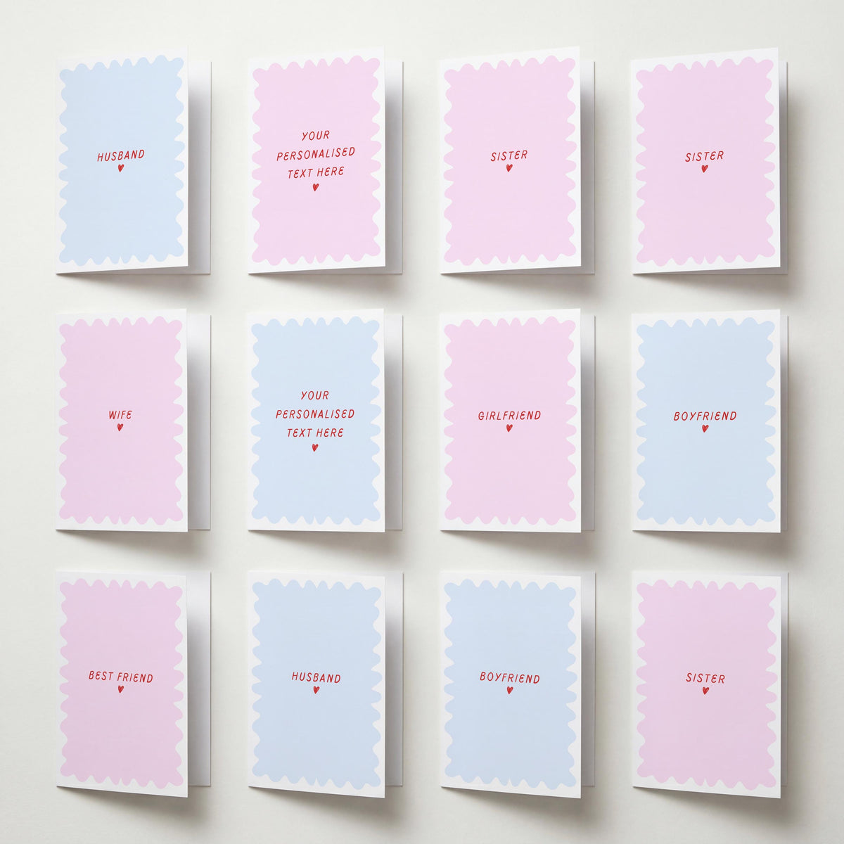 Twelve pastel greeting cards arranged in a 3x4 grid on a light wall. The cards have scalloped edges and are alternating between light pink and light blue, with red text placeholders such as 'HUSBAND', 'SISTER', 'WIFE', 'GIRLFRIEND', 'BOYFRIEND', 'BEST FRIEND', and 'YOUR PERSONALISED TEXT HERE'.